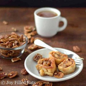 three caramel pecan sticky buns on a plate with a fork set in front of a small dish of pecans and a cup of coffee