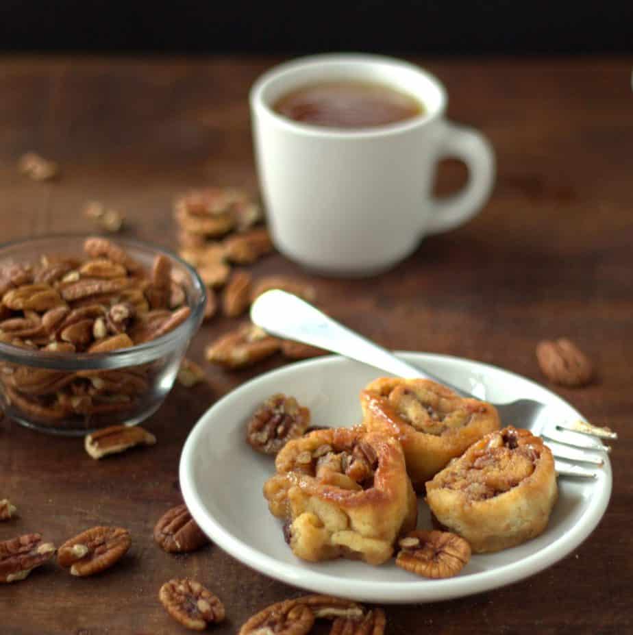 three caramel pecan sticky buns on a white plate with a fork next to a small bowl of pecans