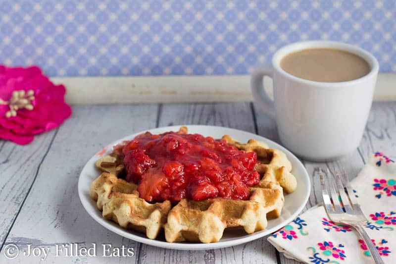 one coconut waffle topped with a fresh strawberry sauce on a white plate next to a white mug of coffee