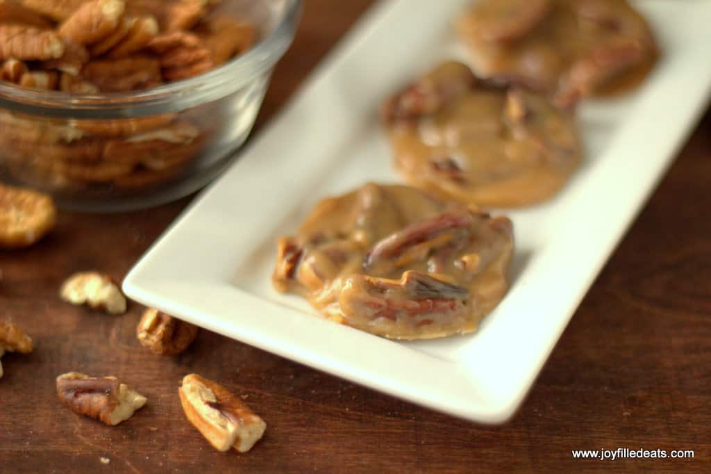 keto pecan pralines lined on a white platter next to a bowl of raw pecans