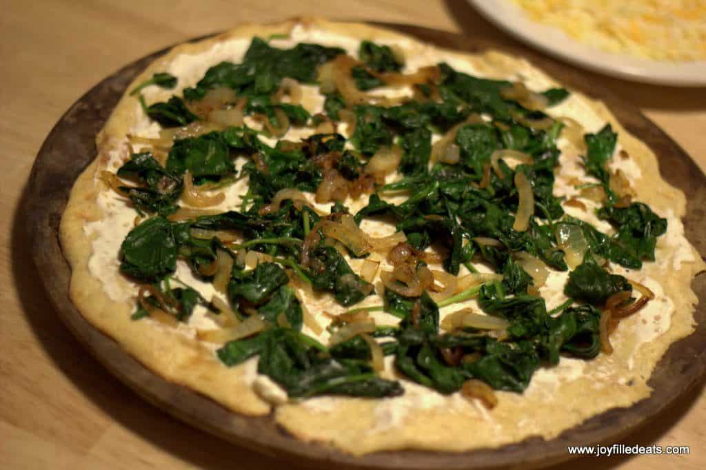 sauteed spinach and onions layered onto keto white pizza placed on a round pizza stone