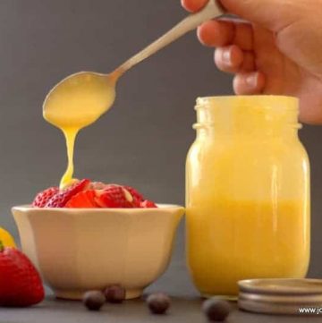 hand using spoon to pour creaming lemon desert sauce onto fruit placed in a white bowl next to mason jar filled with more creamy lemon dessert sauce