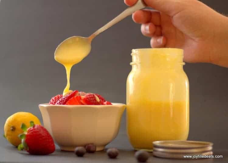 hand using spoon to pour creaming lemon desert sauce onto fruit placed in a white bowl next to mason jar filled with more creamy lemon dessert sauce
