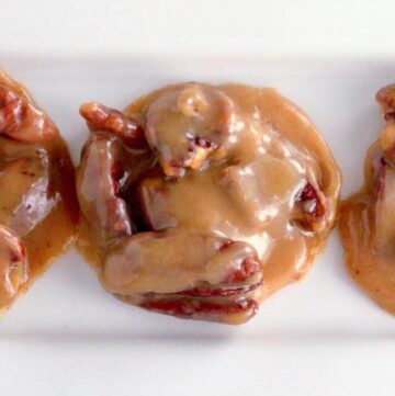 Close up pf 3 Pecan Pralines on a rectangular white plate sitting on a black backdrop with pecans scattered around.