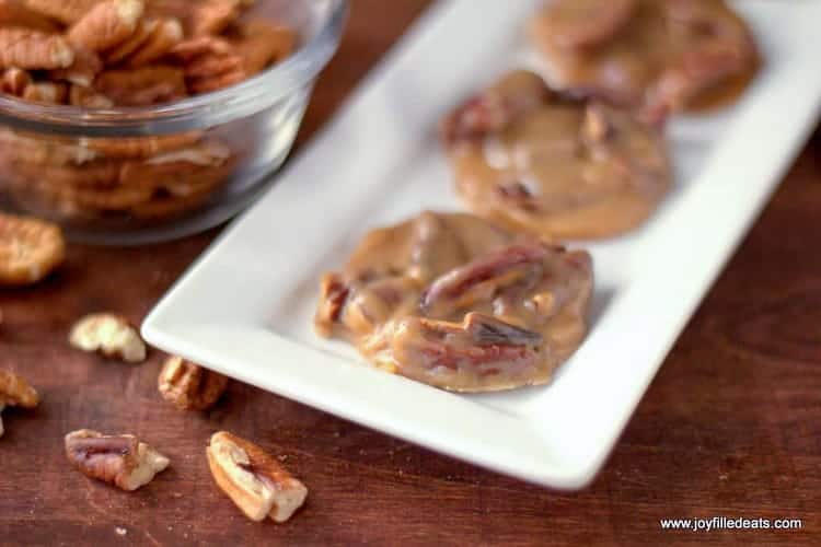 Four Pecan Pralines on a diagonal rectangular white plate sitting on a wood backdrop with pecans in a glass bowl in the corner and scattered around.