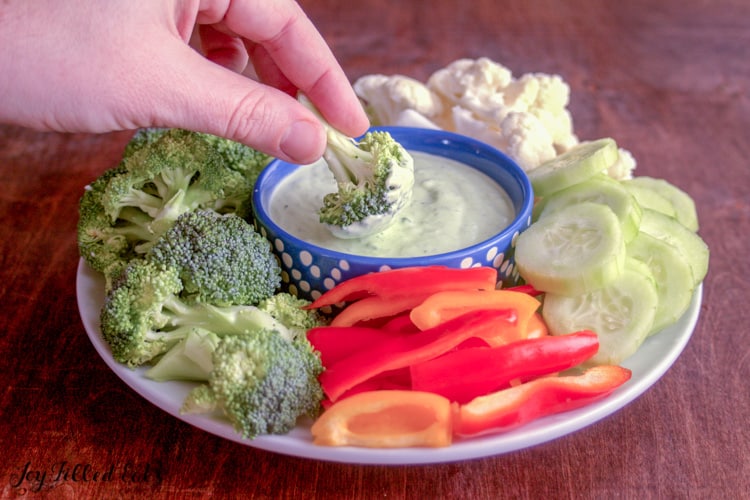 hand dipped broccoli floret into small dish of chive dip placed center on platter surrounded by other vegetables