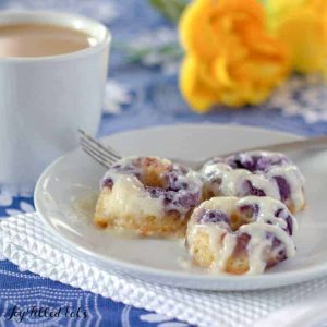 three cream cheese glazed lemon blueberry donuts on a plate with a fork set next to a cup of coffee