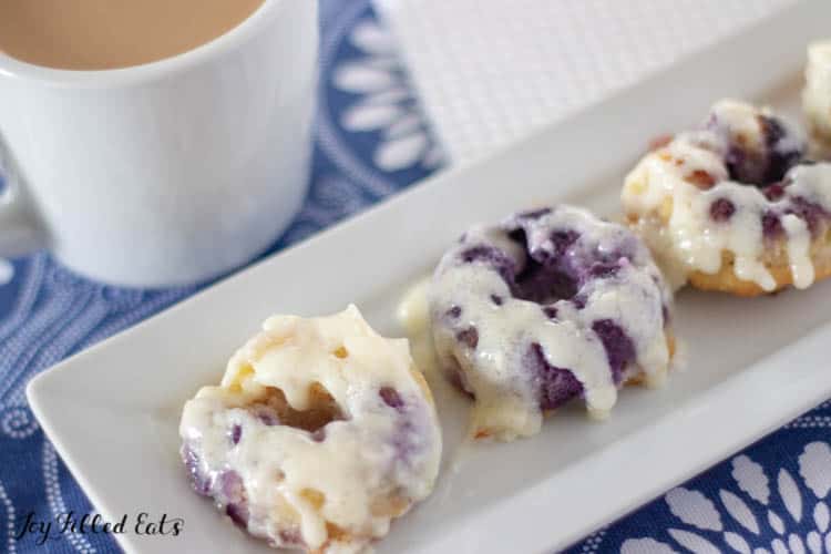 keto lemon blueberry donuts arranged on a platter dripping in a cream cheese glaze next to a cup of coffee