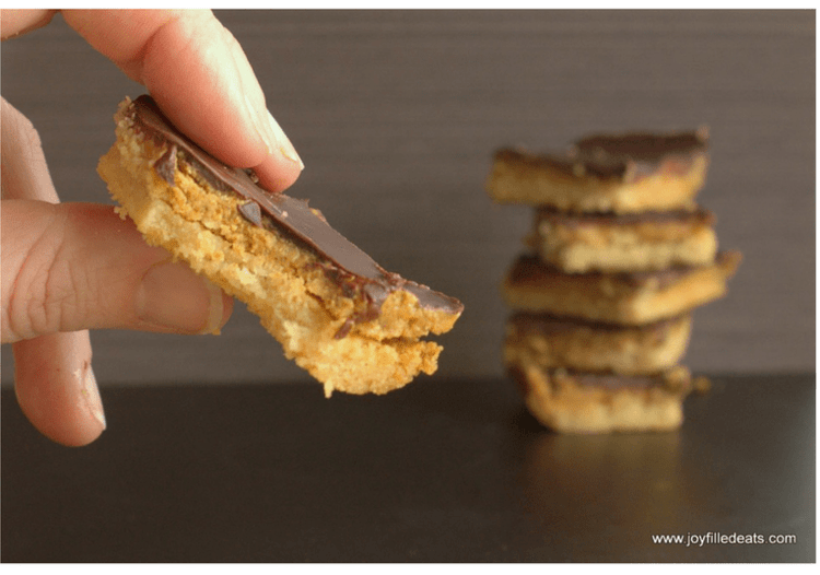 close up of hand holding a homemade tagalong peanut butter cookie bars with bite missing in front of stack of cookies