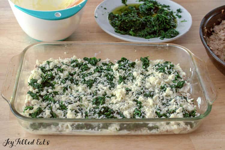 Layered casserole dish with ingredients surround by mixing bowls and plate of spinach