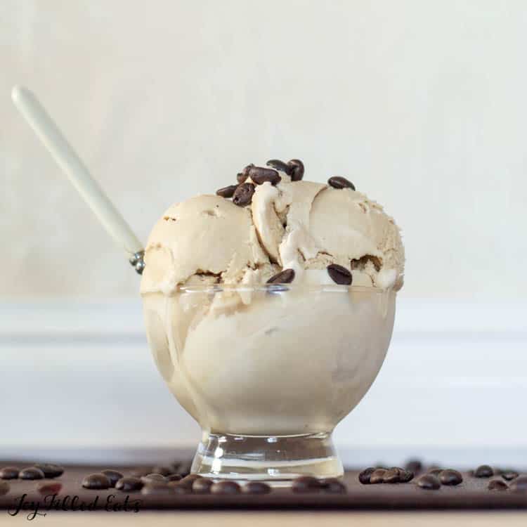 coffee ice cream in a glass bowl with spoon sticking into ice cream topped with coffee beans placed on a table surrounded by scattered coffee beans