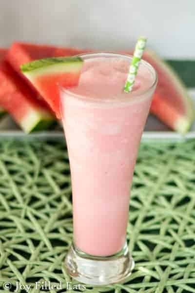 close up on a watermelon smoothie in a tall glass garnished with a fresh watermelon slice and paper straw placed on the glass rim set next to a platter of watermelon slices