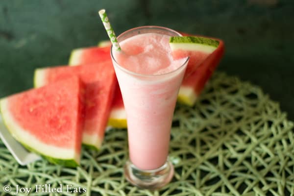 a watermelon smoothie in a tall glass garnished with a fresh watermelon slice placed on the glass rim set next to a platter of watermelon slices