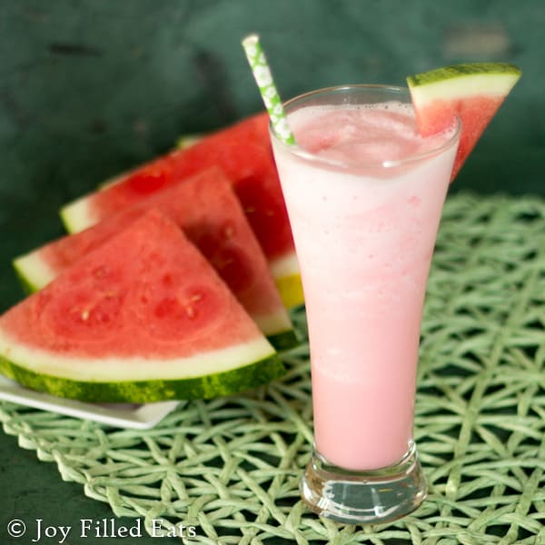 a tall glass filled with a watermelon smoother with a fresh watermelon slice placed on the glass rim set next to a platter of watermelon slices