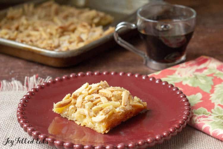 square slice of almond pastry on a red plate next to a cup of coffee and baking dish with remaining pastry