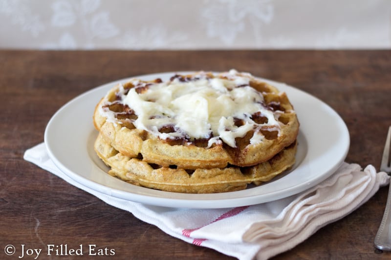short stack of cinnamon roll waffles with cream cheese icing on a white plate set on a white cloth napkin