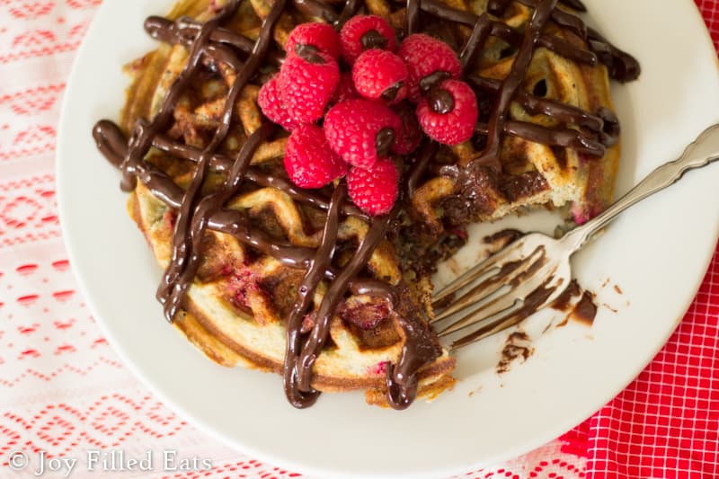 overhead view of chocolate covered raspberry waffles topped with chocolate ganache raspberries with large bite missing and replaced by a fork
