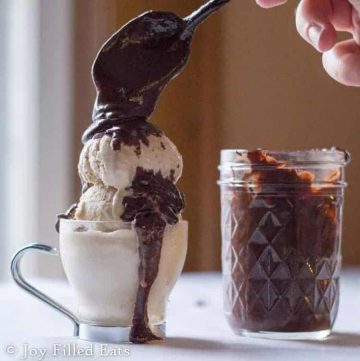 hand holding a spoon full of three ingredient hot fudge being poured onto scoops of ice cream next to a mason jar filled with hot fudge