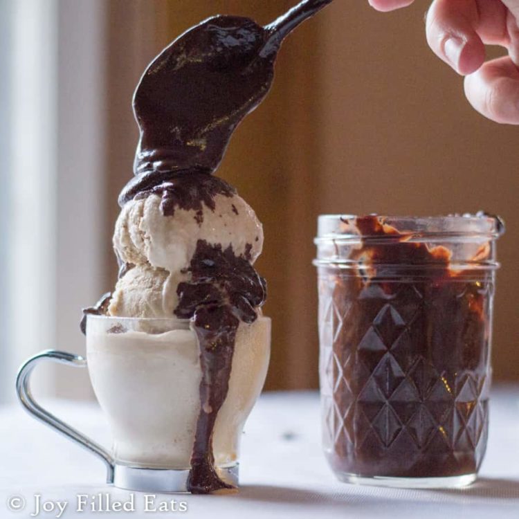 hand holding a spoon full of three ingredient hot fudge being poured onto scoops of ice cream next to a mason jar filled with hot fudge