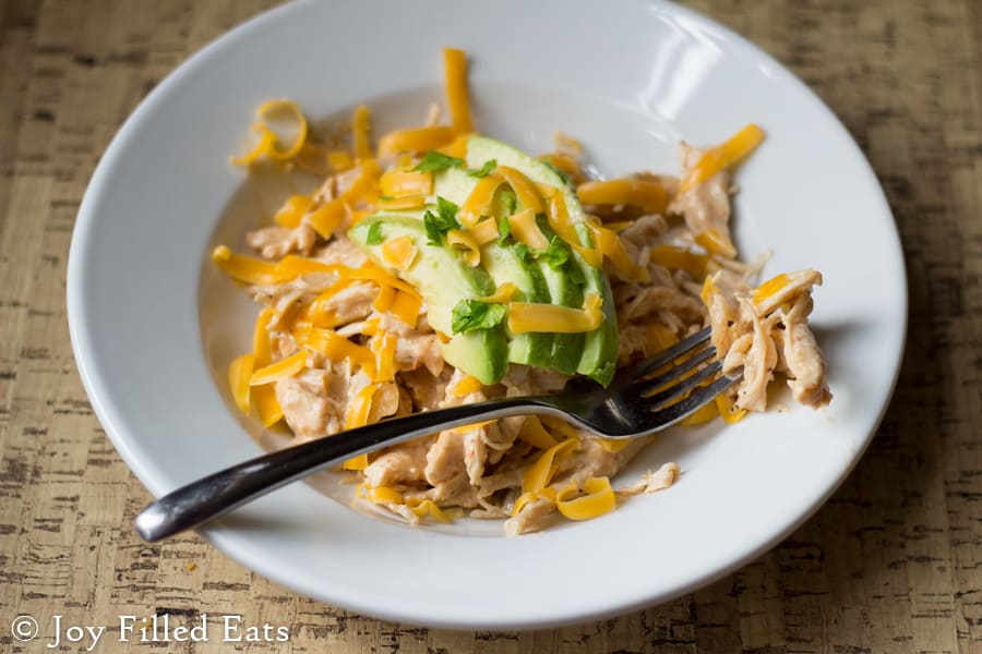 bite of creamy salsa chicken on a fork laying on the rim of a shallow white bowl filled with more creamy salsa chicken and topped with shredded cheese and sliced avocado