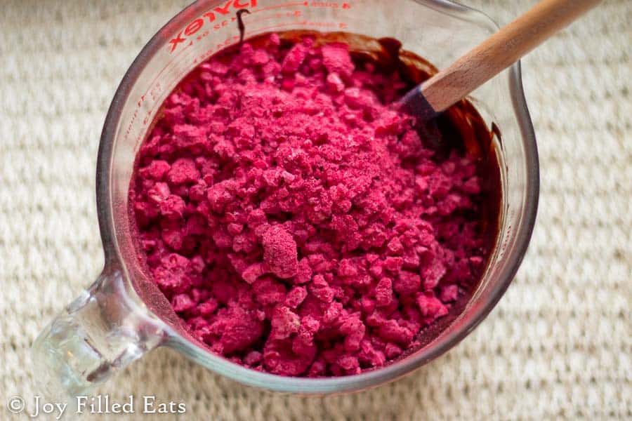 freeze dried raspberries poured into a large mixing bowl of melted dark chocolate with spatula