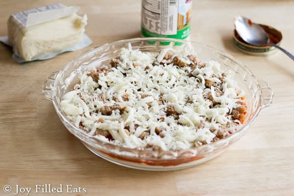 shredded cheese topped on sausage pizza cheese in a glass pie plate