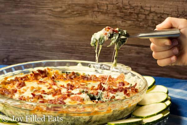 hand holding a small knife lifting a serving of Bacon Caesar Spinach dip for pie plate surrounded by zucchini rounds