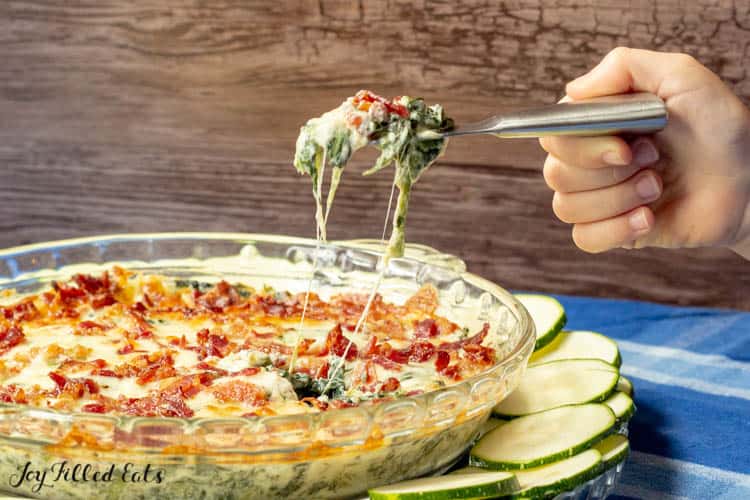 hand scooping a spoonful of bacon spinach dip from pie plate full of baked dip sitting on a blue platter with zucchini slices