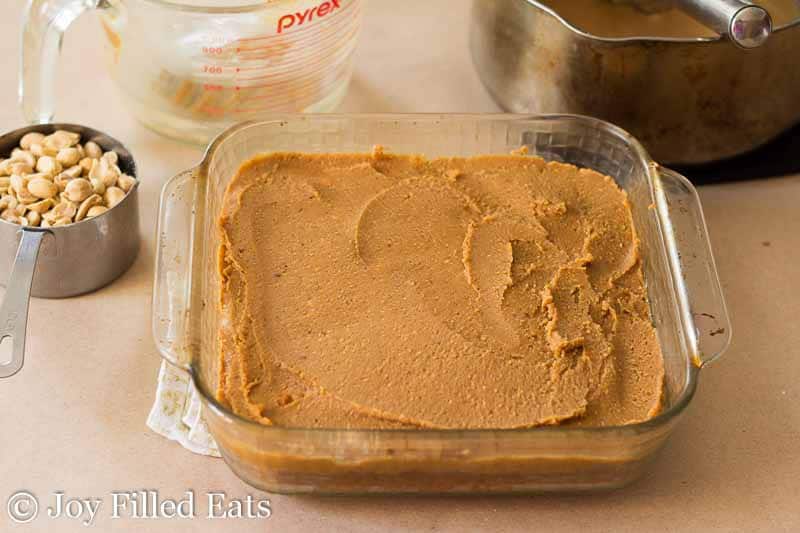 peanut butter filling spread and layered on top of a square baking dish next to a measuring cup of peanuts and saucepan of caramel icing