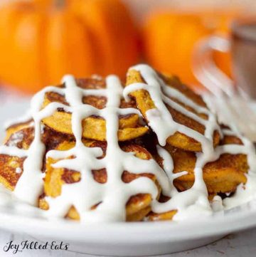 close up of a pile of keto pumpkin pancakes covered in cream cheese icing set in front of a cup of tea and a row of pumpkins