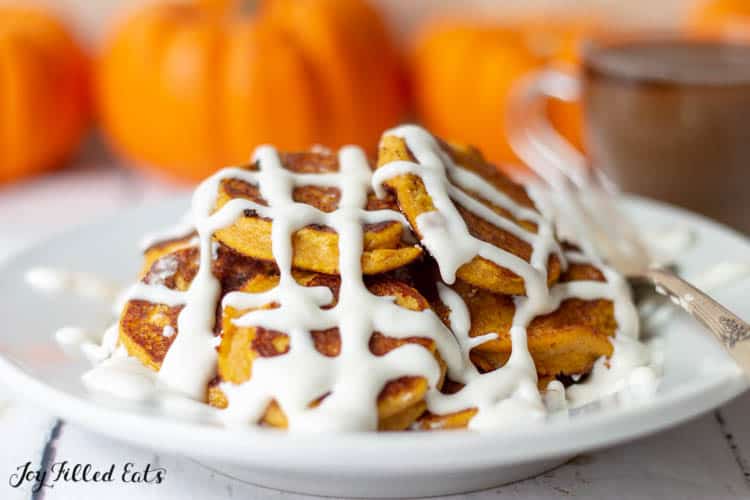 close up of a pile of keto pumpkin pancakes covered in cream cheese icing set in front of a cup of tea and a row of pumpkins