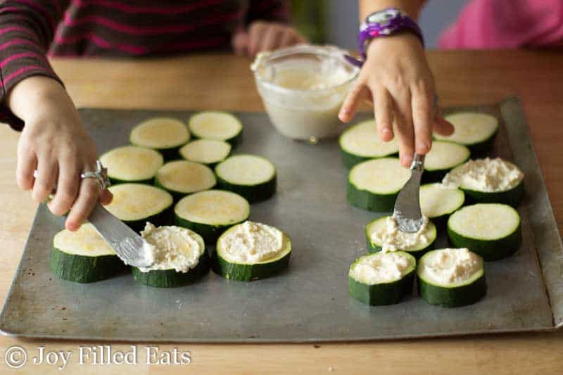 zucchini rounds split in half with two hands working at topping each piece with a Parmesan cheese spread