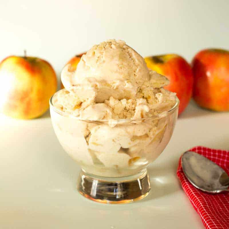 scoop of apple pie ice cream in a glass bowl set in front of a row of apples next to a spoon