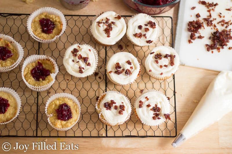 overhead view of cooling rack lined with cranberry bliss cupcakes next to a piping bag of cream cheese frosting and cranberry pieces
