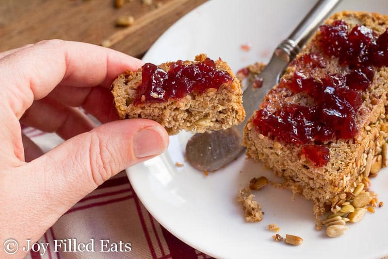 hand holding a piece of sprouted quinoa bread spread with jam above a white plate with butter knife and large slice of jam covered quinoa bread