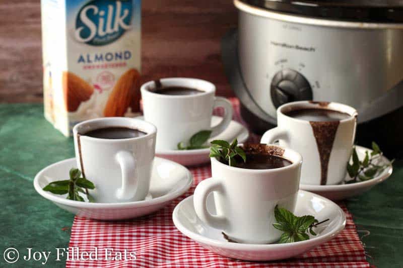 four small white mugs full of crock pot peppermint hot chocolate, all dripping over the sides, and garnished with mint leaves, set in front of a carton of Silk Almond milk and a crock pot