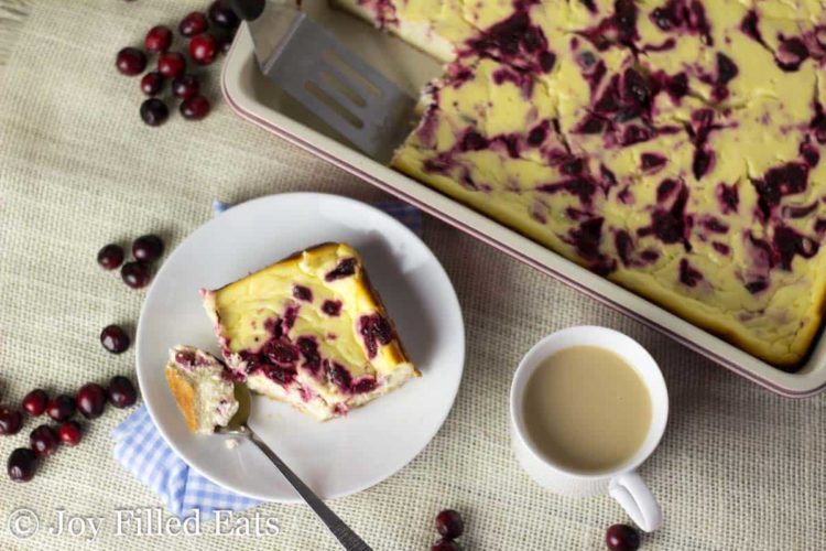 square piece of cranberry swirl cheesecake bars broken up with a spoon next to a mug of coffee and casserole dish with more cranberry cheesecake bars