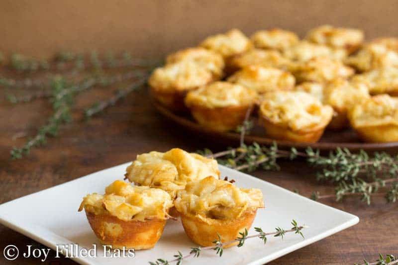 three onion tartlets arranged on a white plate with a sprig of thyme next to a platter of more french onion tartlets