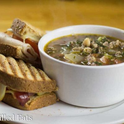 small white bowl of sausage and kale soup on a white plate with a panini sandwich