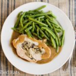 overhead view of white plate with slice of stuffed pork marsala covered in gravy next to a side of green beans placed next to a fork and knife