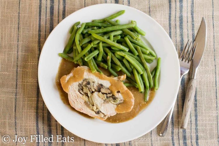 overhead view of white plate with slice of stuffed pork marsala covered in gravy next to a side of green beans placed next to a fork and knife