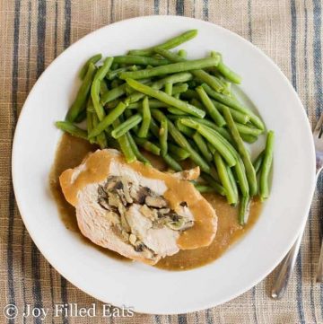 close up on overhead view of white plate with slice of stuffed pork marsala covered in gravy next to a side of green beans placed next to a fork and knife