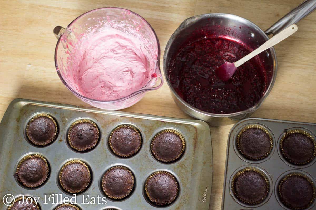 two muffin tins filled with chocolate champagne cupcakes next to a mixing bowl of raspberry icing and a saucepan of raspberry puree