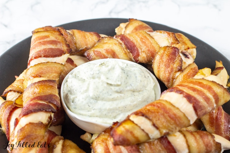 close up on bacon wrapped chicken tenders next to a small dish of ranch dip