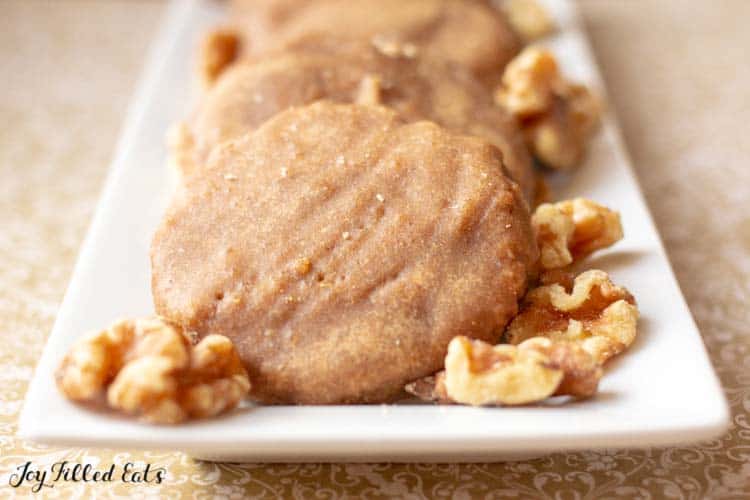 close up on glazed maple walnut cookies fanned out on white, rectangular platter with raw walnuts scattered along side of cookies