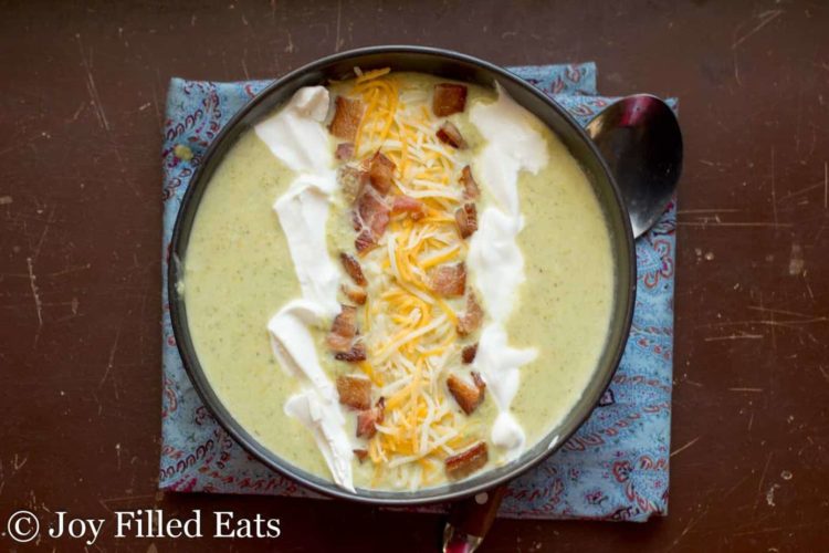 overhead view of loaded broccoli cheddar soup topped with sour cream, bacon bits and shredded cheese, set on a decorative napkin with a spoon