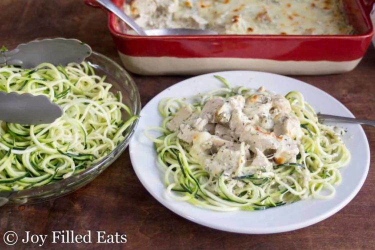 mozzarella & pesto chicken casserole topped on zucchini noodles on a plate set net to a platter of more zucchini noodles and casserole dish