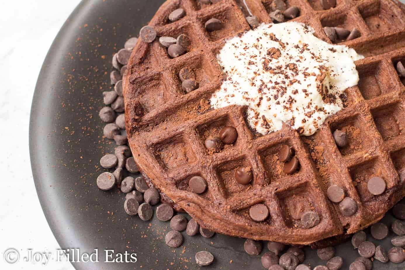chocolate chocolate chip waffle surrounded by chocolate chips and topped with whipped cream on a black plate