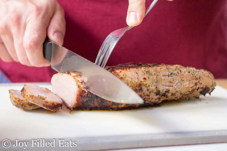 hand using carving knife and fork to slice pork tenderloin on cutting board