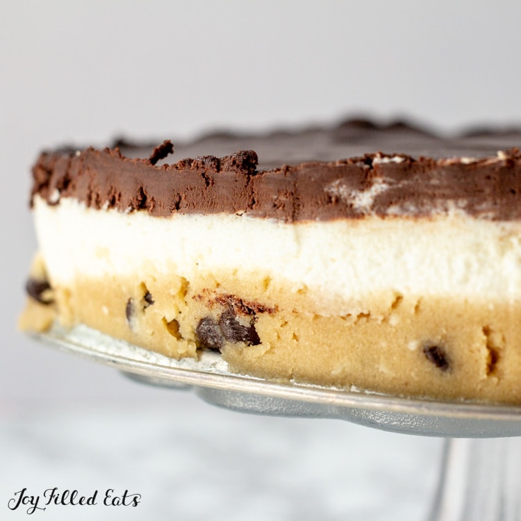 No Bake Cookie Dough Cheesecake on cake platter displaying three layers. Bottom layer of cookie dough, middle layer of cheesecake and top layer of chocolate ganache